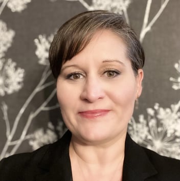Close-up portrait of a woman with short hair, wearing a black blazer, smiling softly against a floral patterned background.
