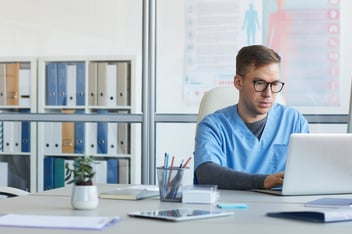 A healthcare professional in light blue scrubs working on a laptop in a modern office, surrounded by office supplies and medical information posters in the background.