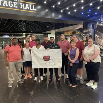 Group photo inside Stage Hall with diverse individuals holding a signed banner for SNU, featuring a modern interior with bright lights.