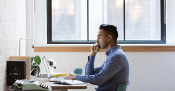 A young man sitting at a desk in a bright, modern workspace, focused on his laptop while deep in thought, with a cup of coffee and a typewriter nearby.