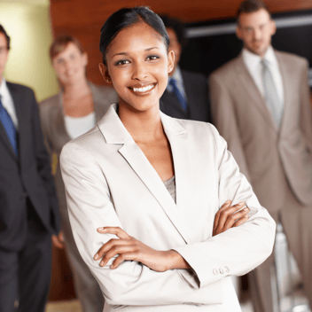 Professional businesswoman smiling confidently with her arms crossed, surrounded by colleagues in a corporate setting.
