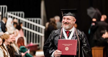 Smiling graduate in a black cap and gown holding a diploma from Southern Nazarene University, celebrating achievement at a formal graduation ceremony with attendees in the background.