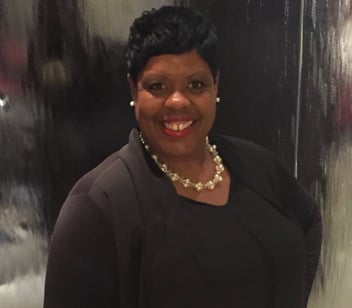 Smiling woman in a black outfit with a pearl necklace posing in front of a shiny dark background.
