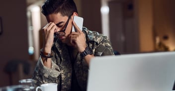 A young man in military uniform sitting at a desk, looking stressed while talking on the phone, with a laptop and coffee cup in the foreground.