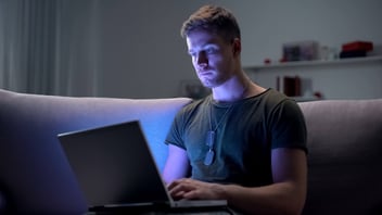 A young man using a laptop at night, illuminated by the screen's blue light while sitting on a couch.