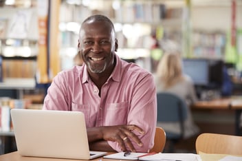 Smiling older man sitting at a laptop in a library, wearing a pink shirt, with notebook and pen in front of him, conveying a sense of engagement and positivity.
