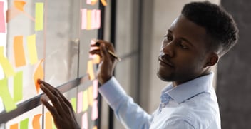 A focused professional examining colorful sticky notes on a glass wall, representing brainstorming and project planning in a collaborative office environment.