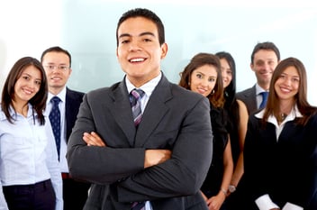 A diverse group of professionals smiling together in a modern office setting, with a confident man in a suit standing front and center, showcasing teamwork and collaboration.