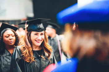Smiling graduate wearing a cap and gown during a graduation ceremony, with fellow graduates visible in the background.