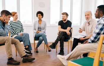 A diverse group of six people engaged in a discussion or therapy session, seated in a circle in a bright, modern room, with a mix of expressions and styles.