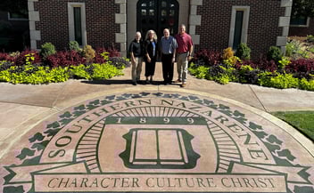 Group of four individuals standing in front of the Southern Nazarene University emblem surrounded by vibrant landscaping.