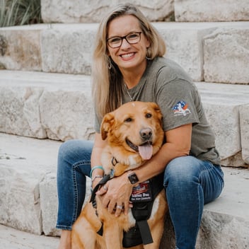 Smiling woman sitting on stone steps, cuddling a golden retriever service dog wearing a vest, showcasing a warm and friendly bond.