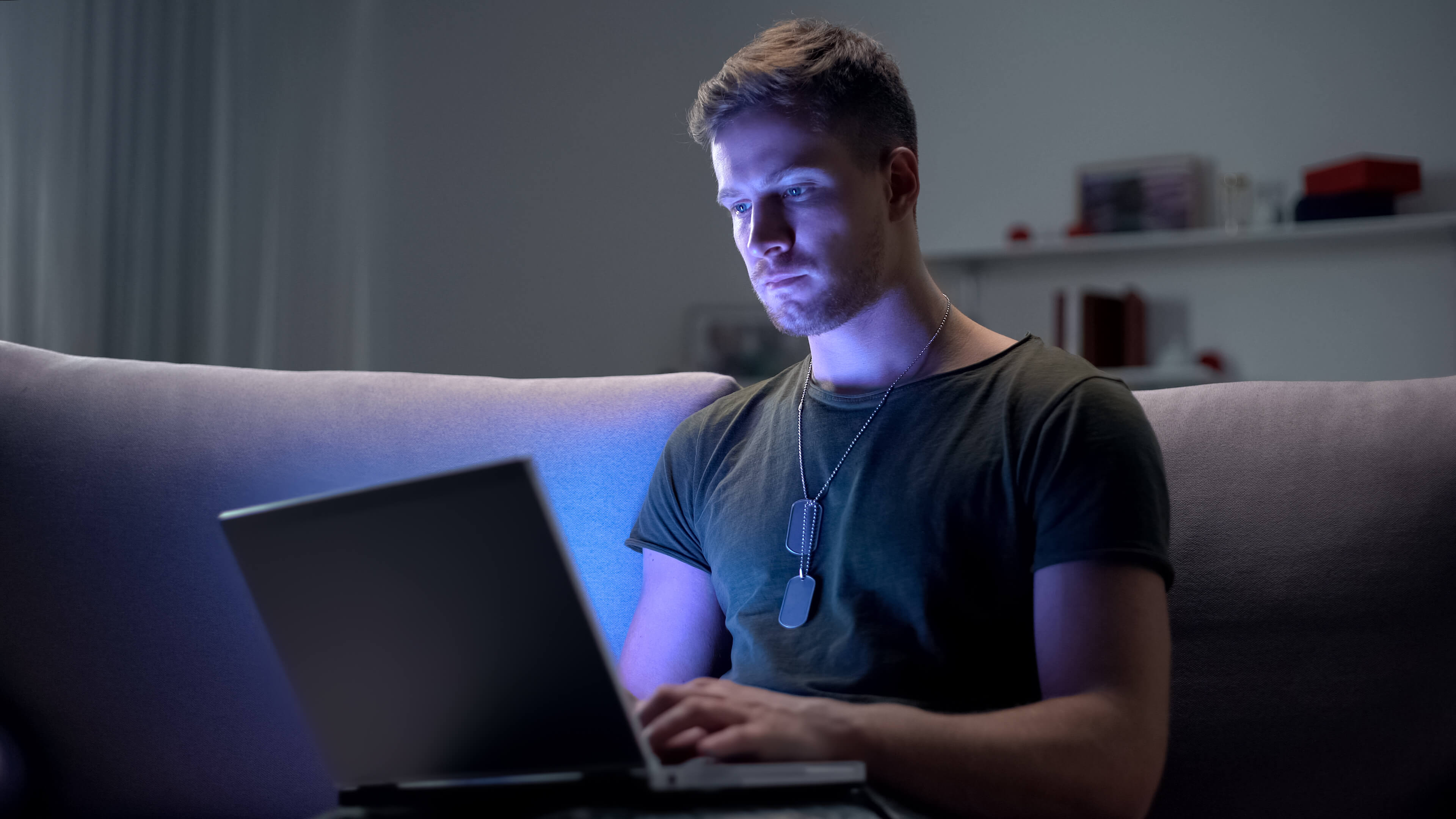 A young man using a laptop at night, illuminated by the screen's blue light while sitting on a couch.