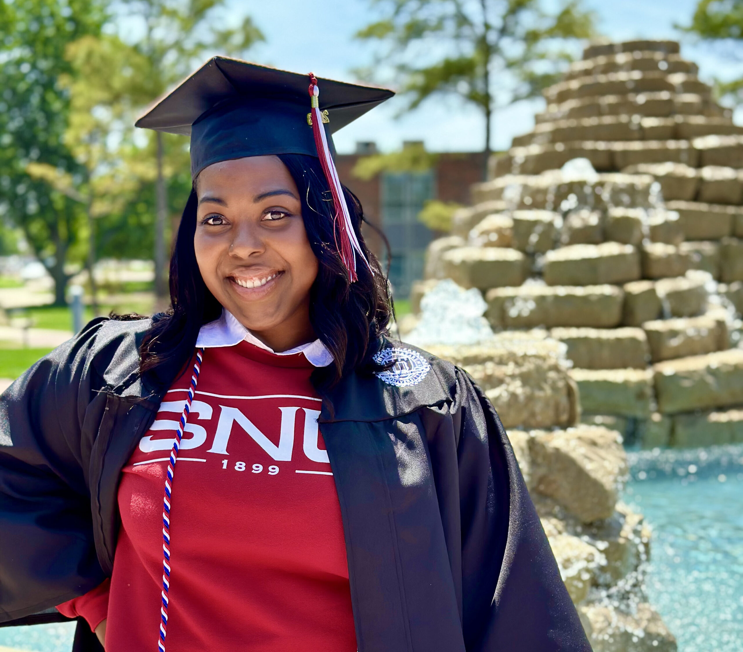 Tiffany Taylor standing in front of a fountain wearing an SNU shirt and graduation regalia